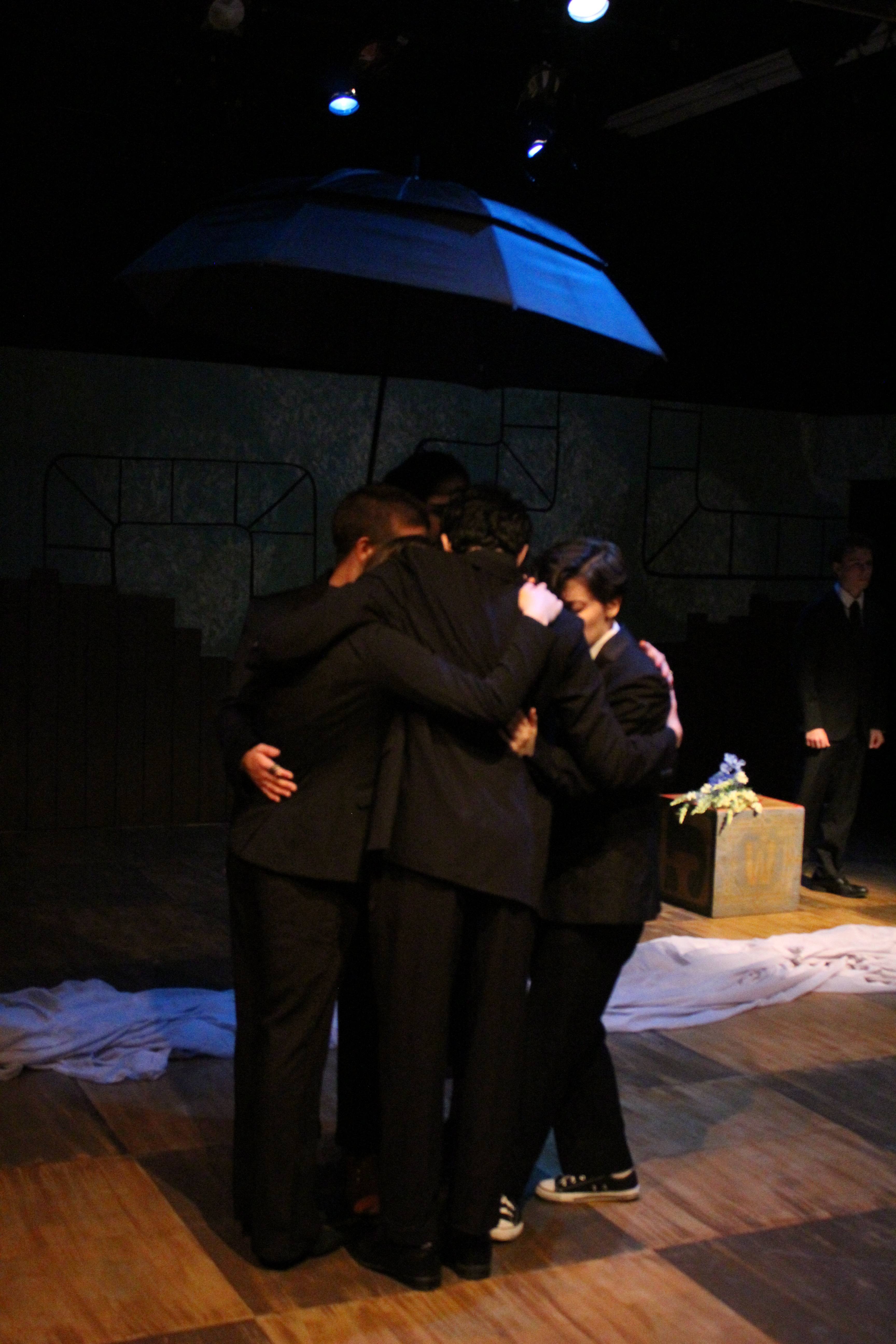 Falsettos - Group embrace under umbrella at funeral