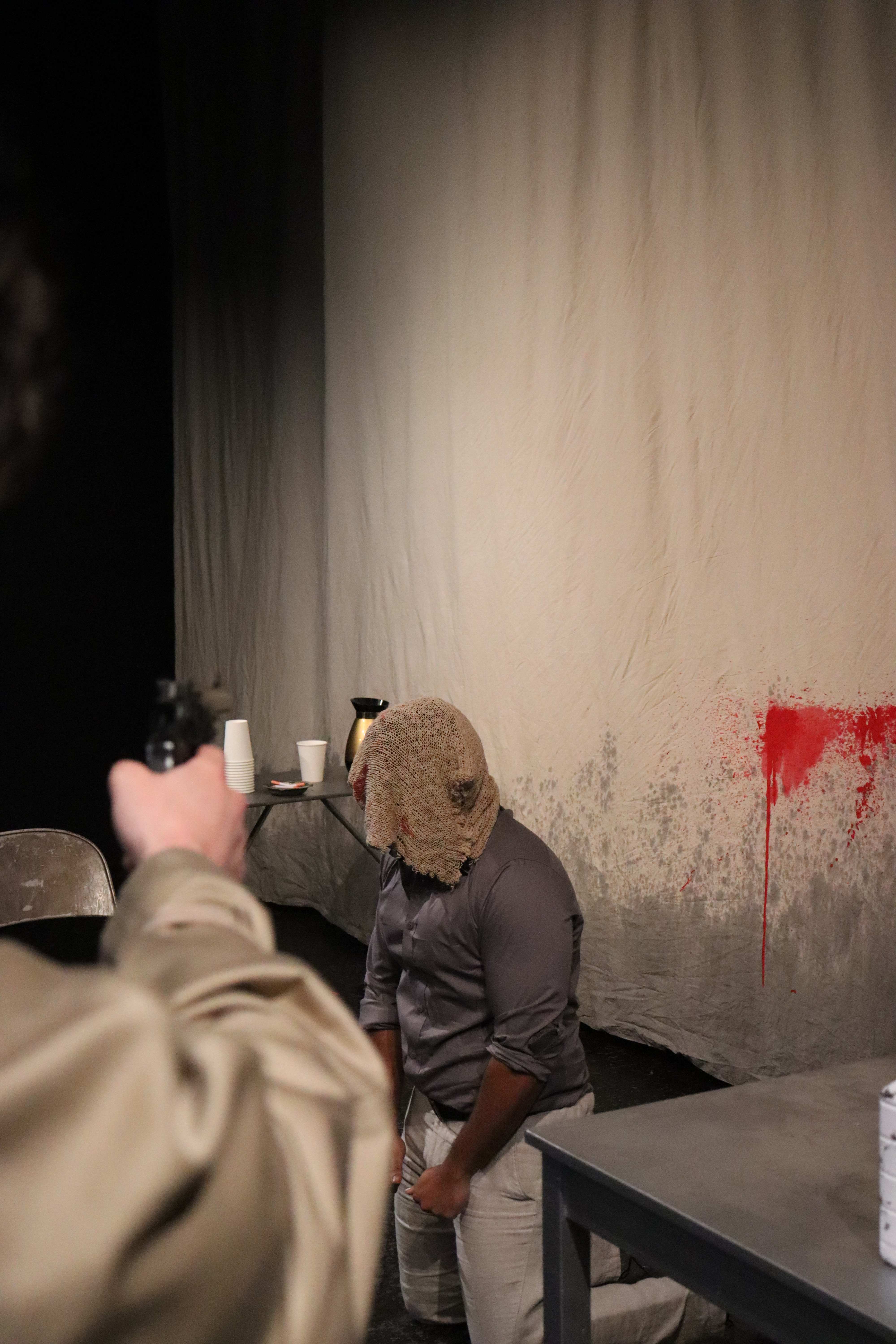 The Pillowman - Man with burlap hood at gunpoint in interrogation room